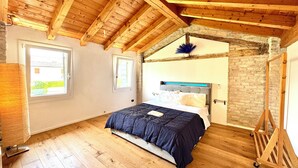 Quarto com teto de madeira, cama com colcha azul escura, parede de pedra e janela com vista para vegetação.