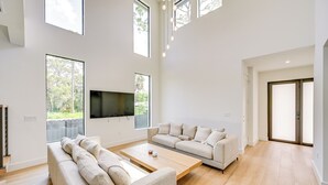 A spacious living room with a large flat-screen TV, two sofas, a coffee table, and large windows offering a view of greenery. - 3 Mi to Naples Beach: Luxurious Home w/ Pool! (Naples)