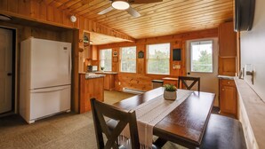 Interior de cabana de madeira com mesa de jantar, cadeiras, geladeira, ventilador de teto e cozinha.