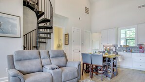 Una sala de estar moderna con un sofá gris, un comedor con mesa y sillas, y una cocina con armarios blancos y un ventanal.