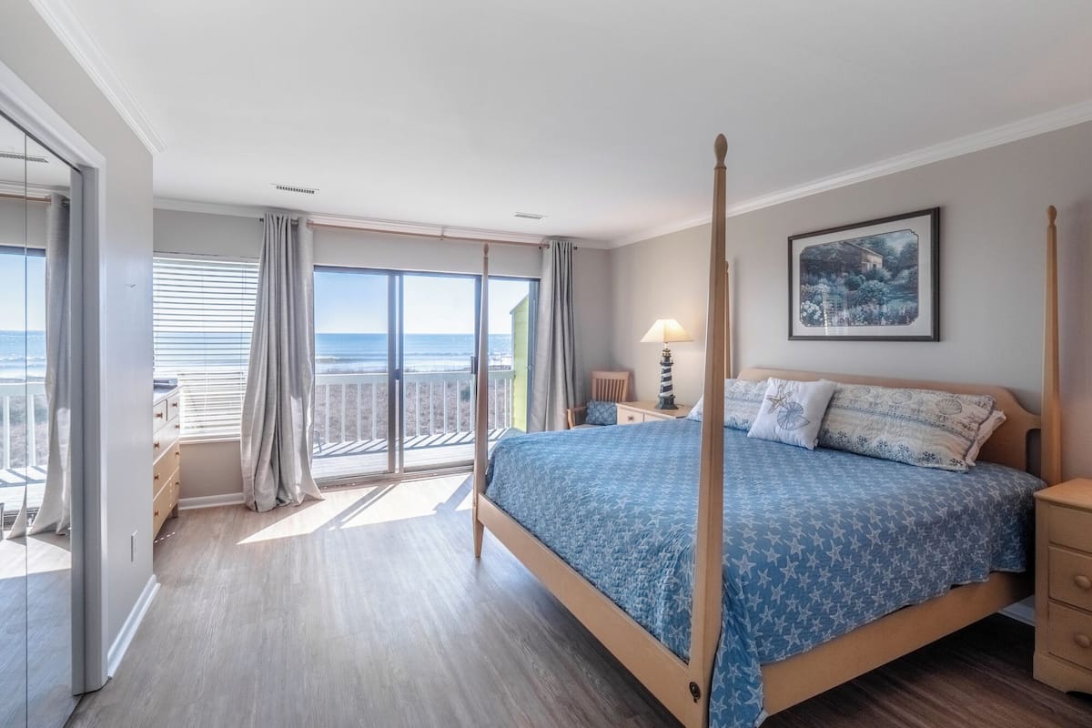 A bedroom with a four-poster bed, a balcony with a view of the beach, and a painting on the wall.