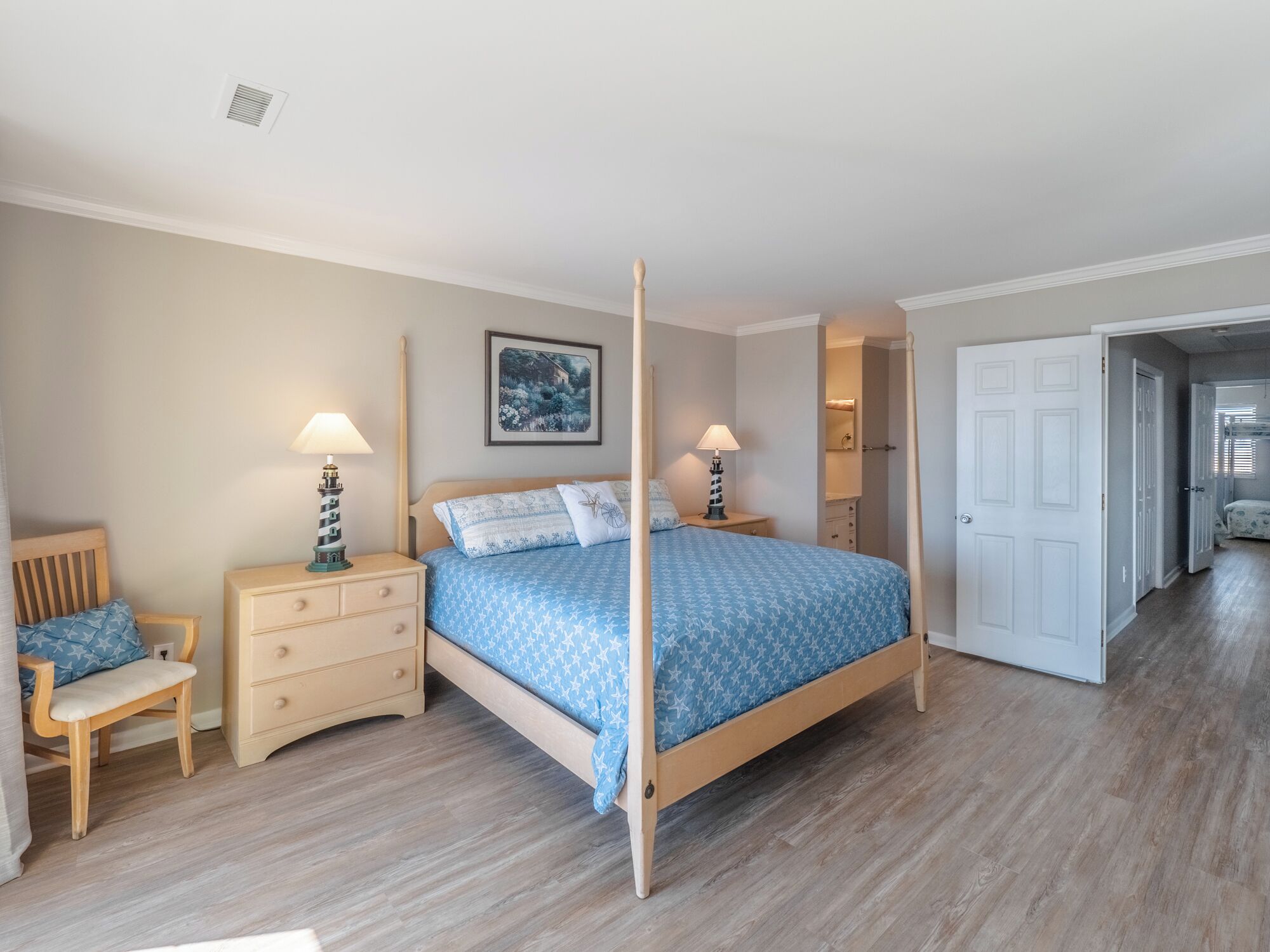 A bedroom with a four-poster bed, a nightstand, a chair, and a door leading to another room.
