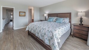A bedroom with a wooden bed, two bedside tables with lamps, a nightstand, and a hallway leading to another room. - Here Comes The Sun by Carolina Retreats (Carolina Beach)