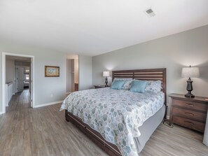 A bedroom with a wooden bed, two bedside tables with lamps, a nightstand, and a hallway leading to another room.