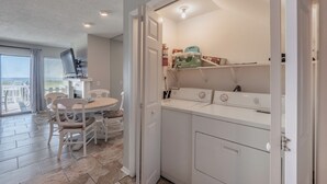 A compact kitchen with white appliances, a dining area with a table and chairs, and a view through a large window. - Here Comes The Sun by Carolina Retreats (Carolina Beach)