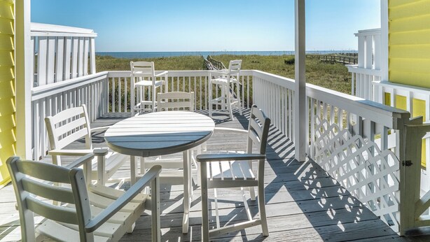 A wooden deck with white railings and a table with chairs, overlooking a beach. - Here Comes The Sun by Carolina Retreats (Carolina Beach)