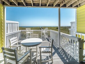 Eine Holzterrasse mit weißen Geländern und einem Tisch mit Stühlen, mit Blick auf einen Strand.