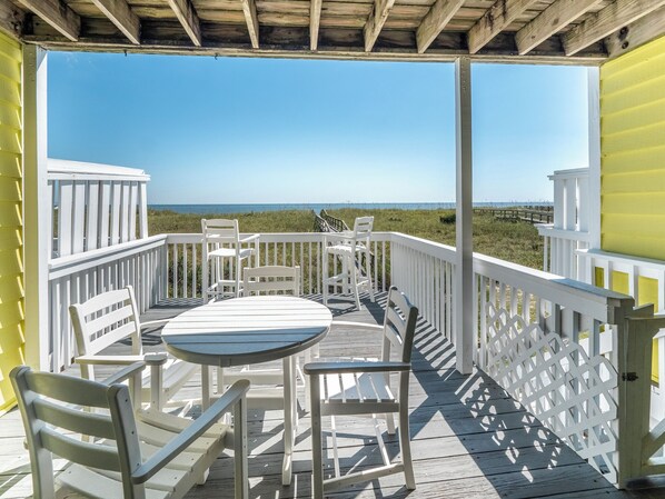 A wooden deck with white railings and a table with chairs, overlooking a beach.
