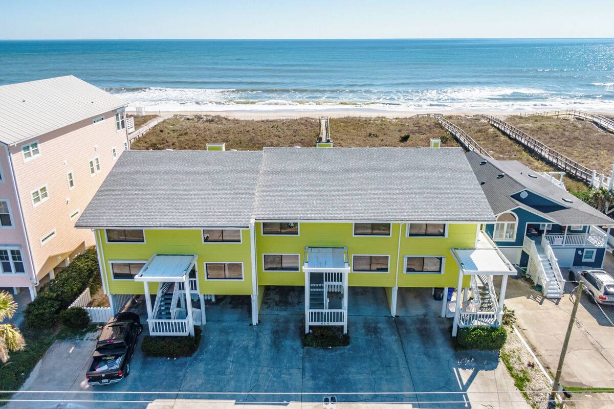 Un edificio de varios pisos con techo plano, cerca de una playa con un paseo marítimo de madera.