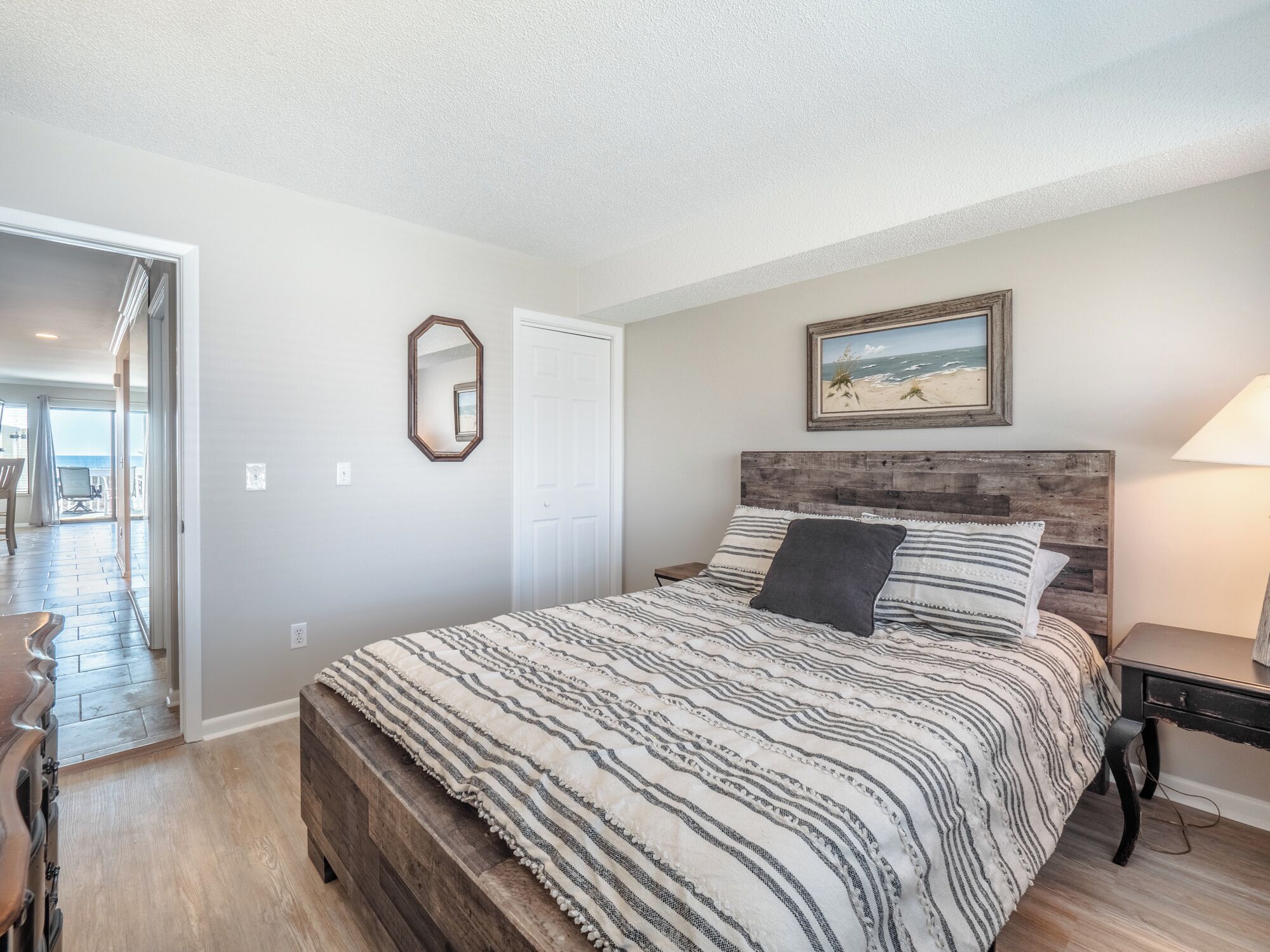 A bedroom with a large bed, a wooden headboard, a painting of a beach, and a door leading to another room.