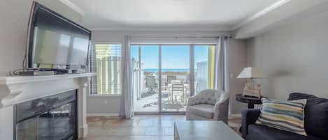 A modern living room with a fireplace, a flat-screen TV, a sofa, a coffee table, and a balcony with a view of the beach.