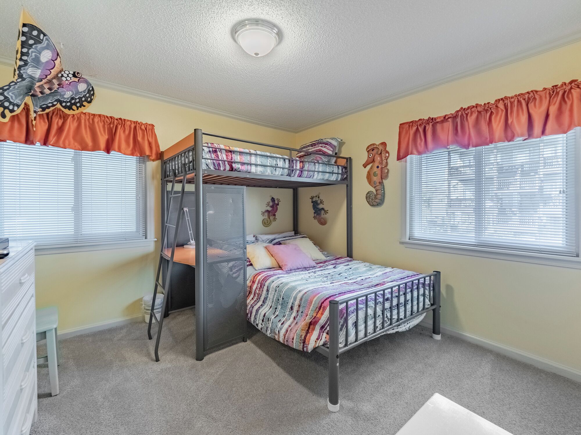 A bunk bed room with a ladder, a desk, and two windows with orange curtains.