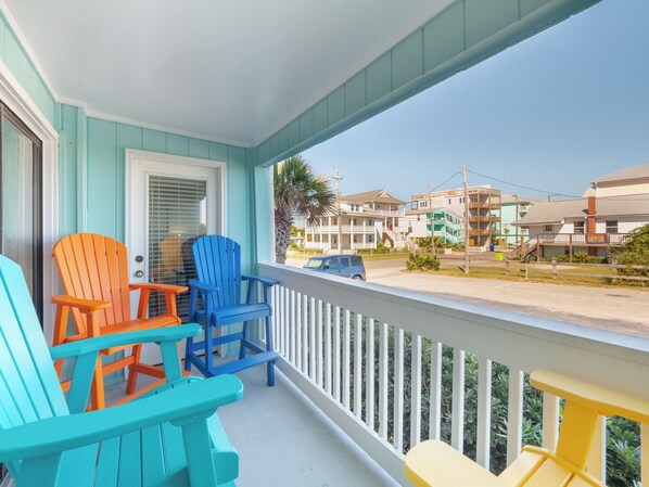 A porch with colorful Adirondack chairs, a white railing, and a view of a street with buildings. - A Place At The Beach #1a- Badabing by Carolina Retreats (Carolina Beach)