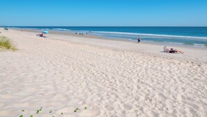 Une vaste plage de sable, parsemée d’empreintes et de quelques plantes clairsemées, un ciel bleu dégagé et des vagues océaniques douces.