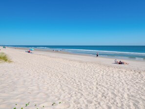 A wide sandy beach with footprints and sparse vegetation, a clear blue sky, and calm ocean waves. - A Place At The Beach #1a- Badabing by Carolina Retreats (Carolina Beach)
