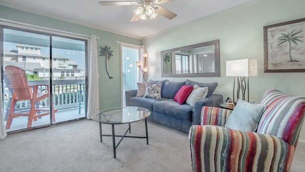 Un salon avec une table en verre, un ventilateur de plafond, un canapé, un fauteuil coloré et un balcon avec une chaise.