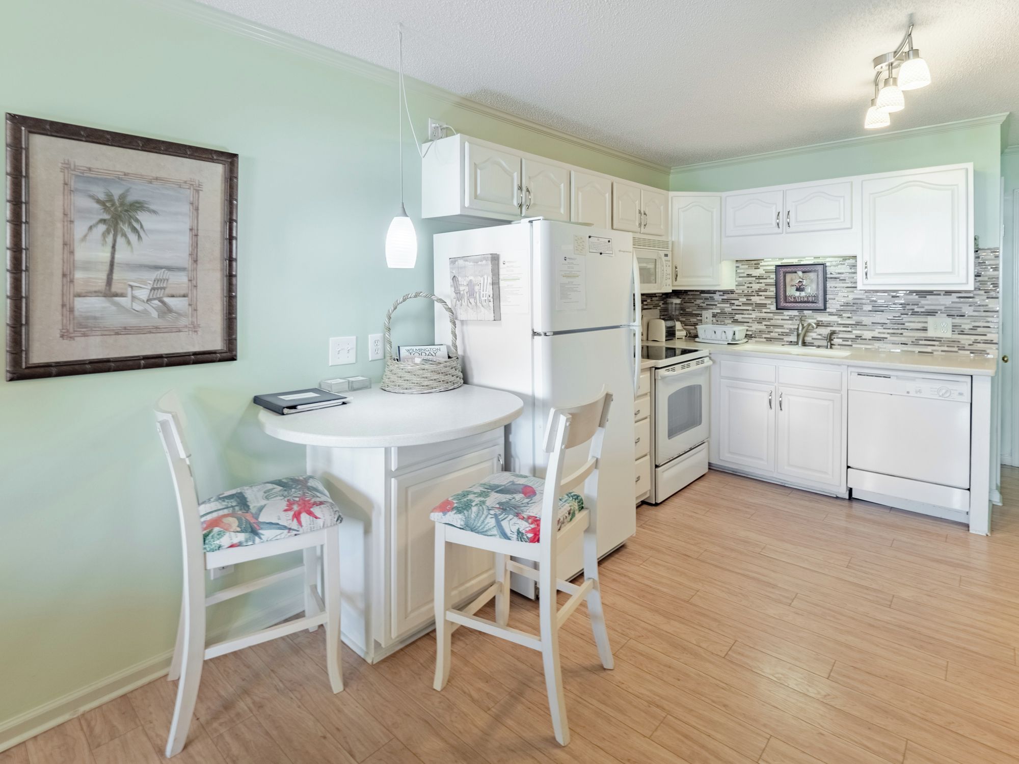 A kitchen with white cabinets, a refrigerator, a stove, and a sink. There is a dining table with chairs and a hanging light fixture.