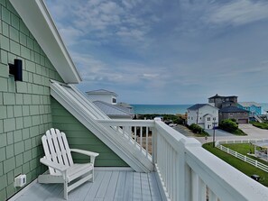 Un balcón con una silla Adirondack blanca, vista al océano y una hilera de casas al fondo.