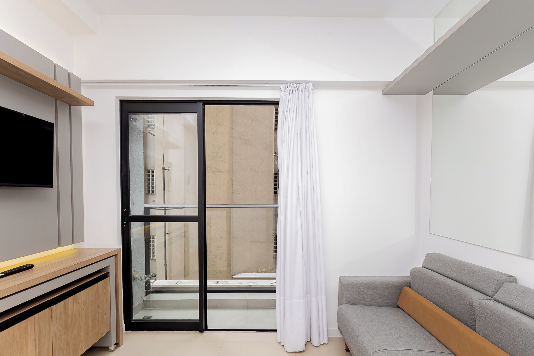 Un salon moderne avec un canapé gris, un meuble en bois, un téléviseur à écran plat et une porte-fenêtre coulissante donnant sur un espace extérieur.
