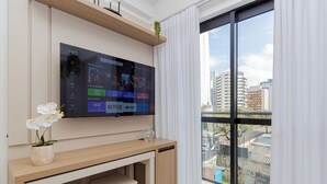 A modern living room with a large flat-screen TV mounted on the wall, a wooden desk with a potted orchid, and a view of a cityscape through large windows. - Start - Gonzaga Stays (Curitiba)