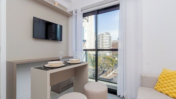 A modern apartment with a dining area, a flat-screen TV, a large window with a city view, and a yellow cushion on a sofa. - Start - Gonzaga Stays (Curitiba)
