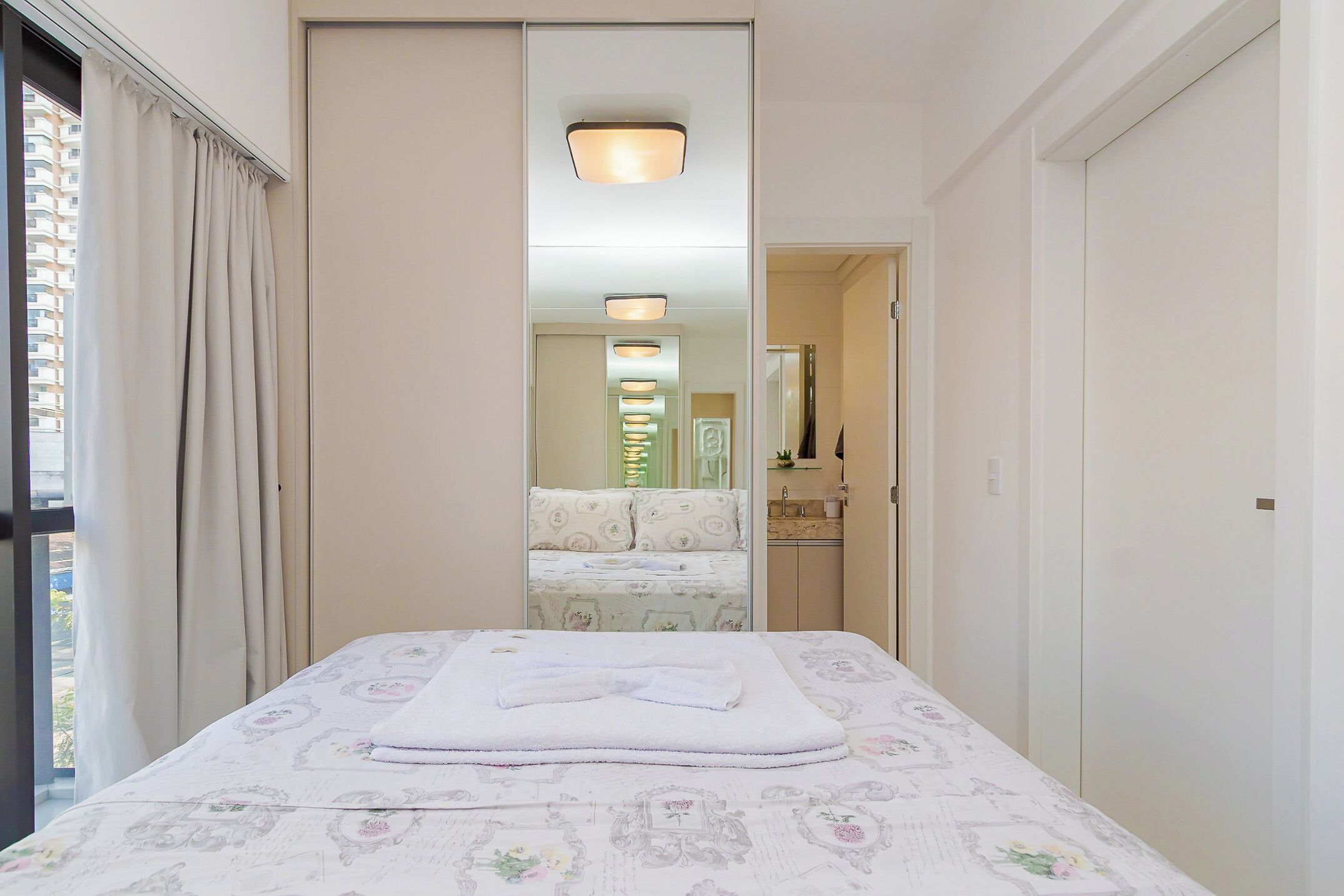 A hotel room with a bed, a mirror, a window with curtains, and a bathroom visible through the mirror.