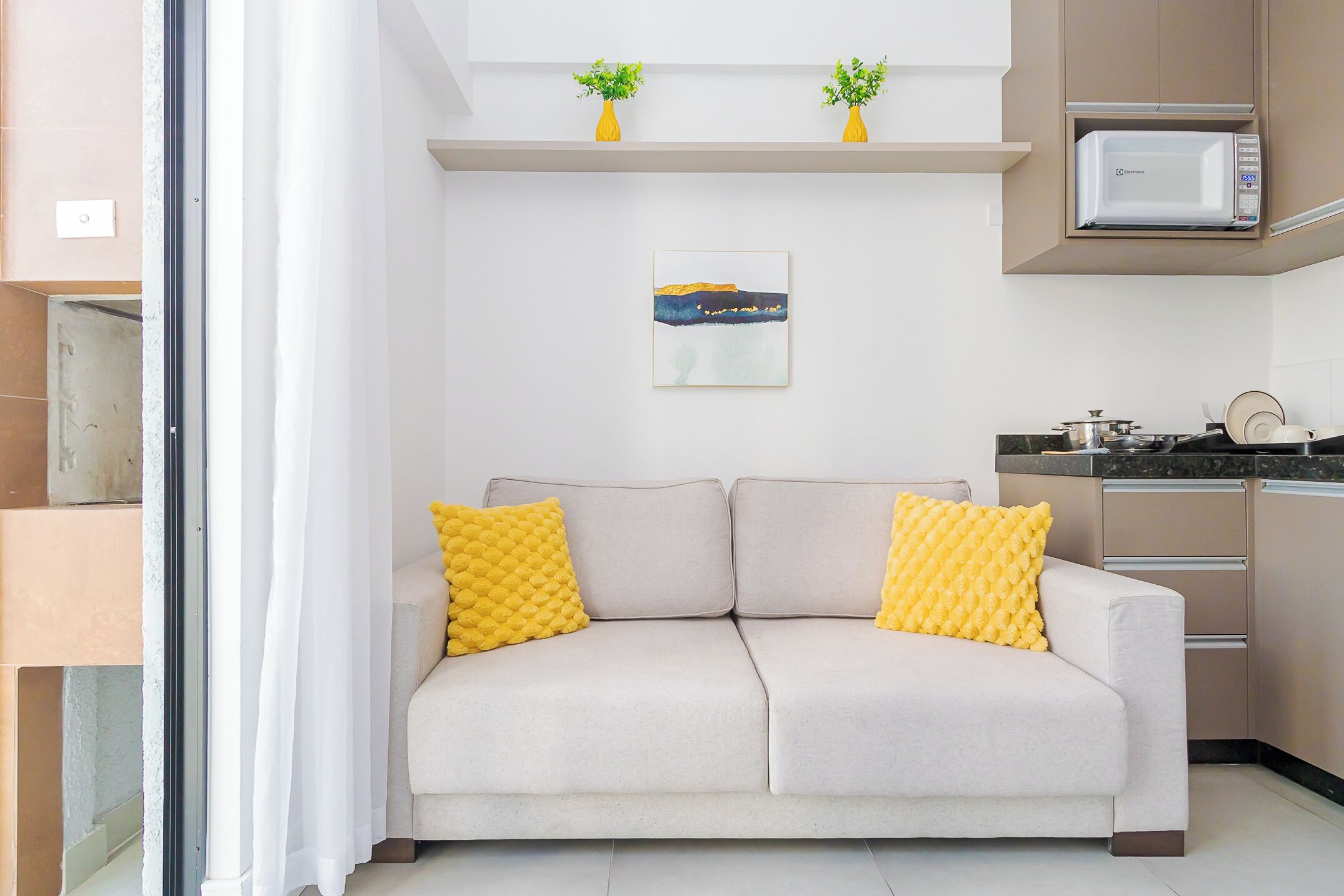 A modern living room with a sofa, yellow cushions, a painting, and a kitchen area with a microwave.