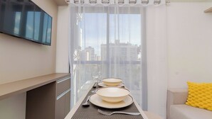 A modern kitchen with a flat-screen TV, a dining table set with plates and cutlery, and a view of the city through sheer curtains. - Start - Gonzaga Stays (Curitiba)