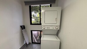 Un cuarto de lavado con lavadora y secadora apiladas verticalmente, una ventana con vistas a la vegetación y una plancha apoyada contra la pared.