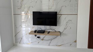 Un baño moderno con pared de mármol, un televisor montado y un estante de madera con dispositivos electrónicos.