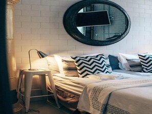 Habitación de hotel ordenada con una cama, un taburete pequeño, un espejo redondo y un televisor fijado a la pared.