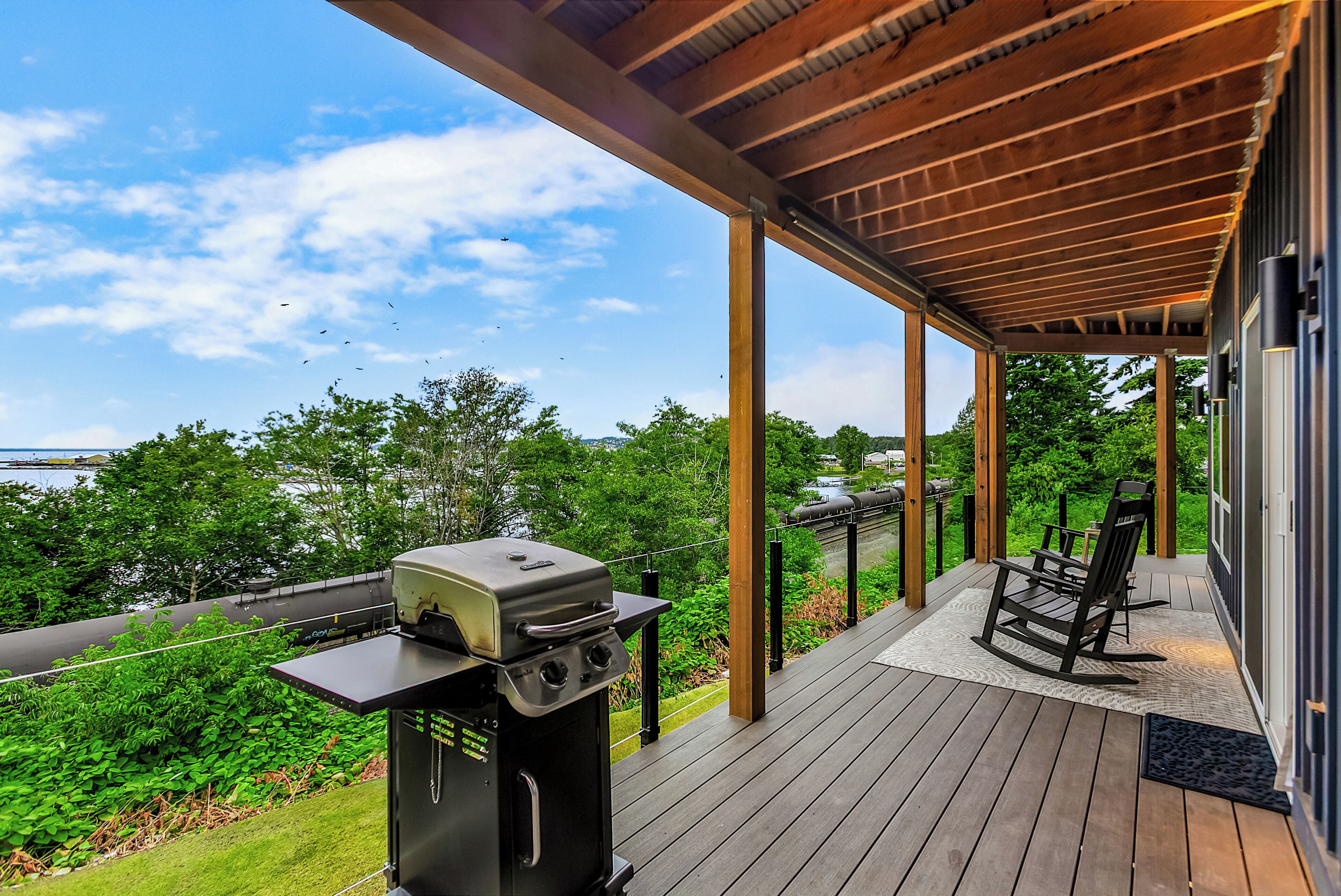 A covered deck with a grill, outdoor seating, and a view of the surrounding greenery and water.