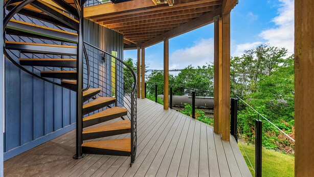 Una escalera de caracol con escalones de madera y barandillas de metal, que conduce a una terraza cubierta con vistas a follaje y un tren al fondo.