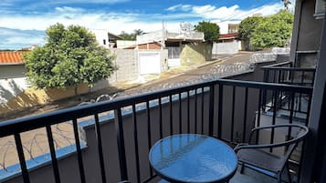 A balcony with a glass table and two wicker chairs, overlooking a residential street with houses and a clear sky.