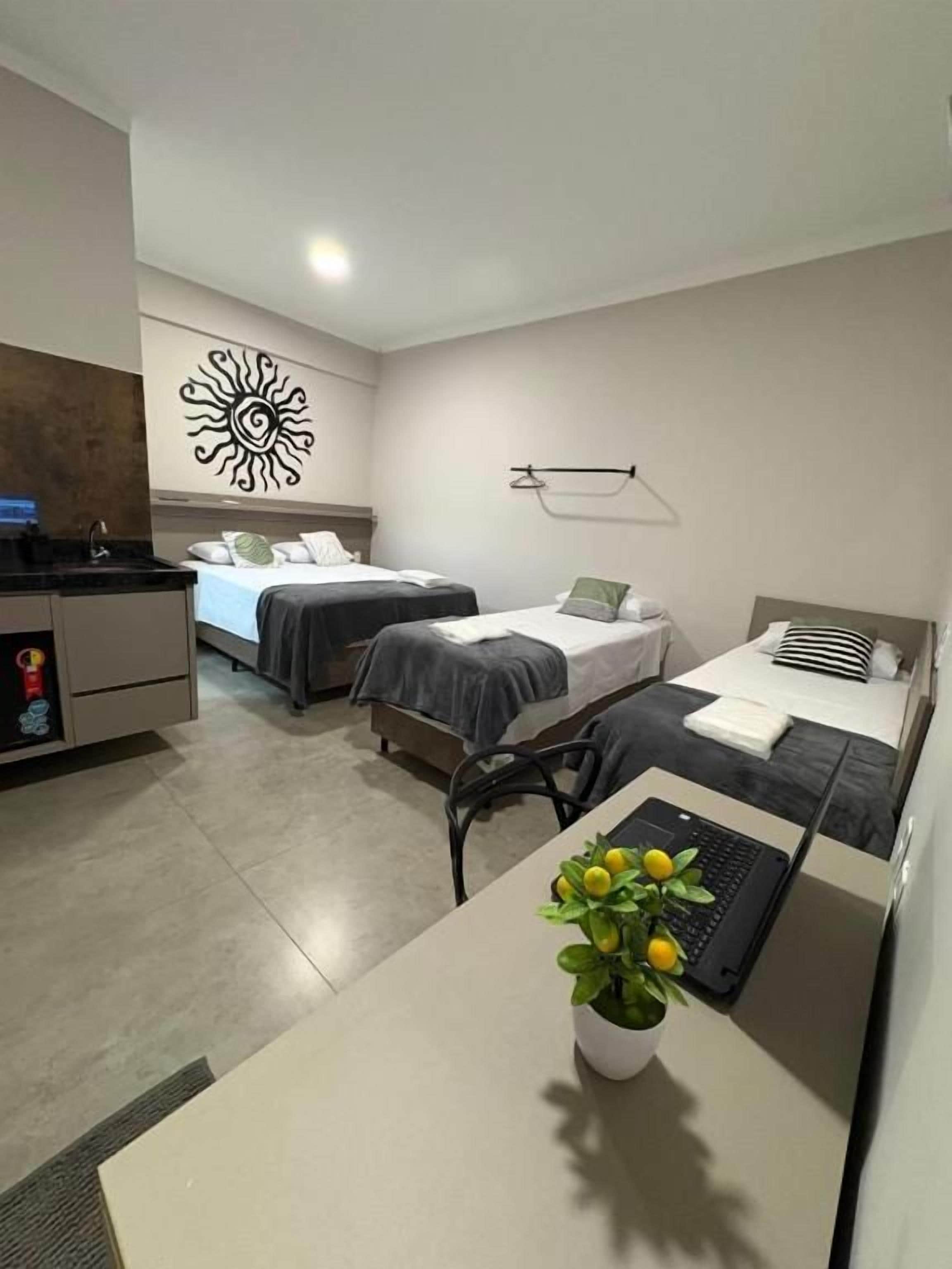 Quarto de hotel com duas camas, uma escrivaninha com um laptop, uma pequena planta e um cabideiro fixado na parede.