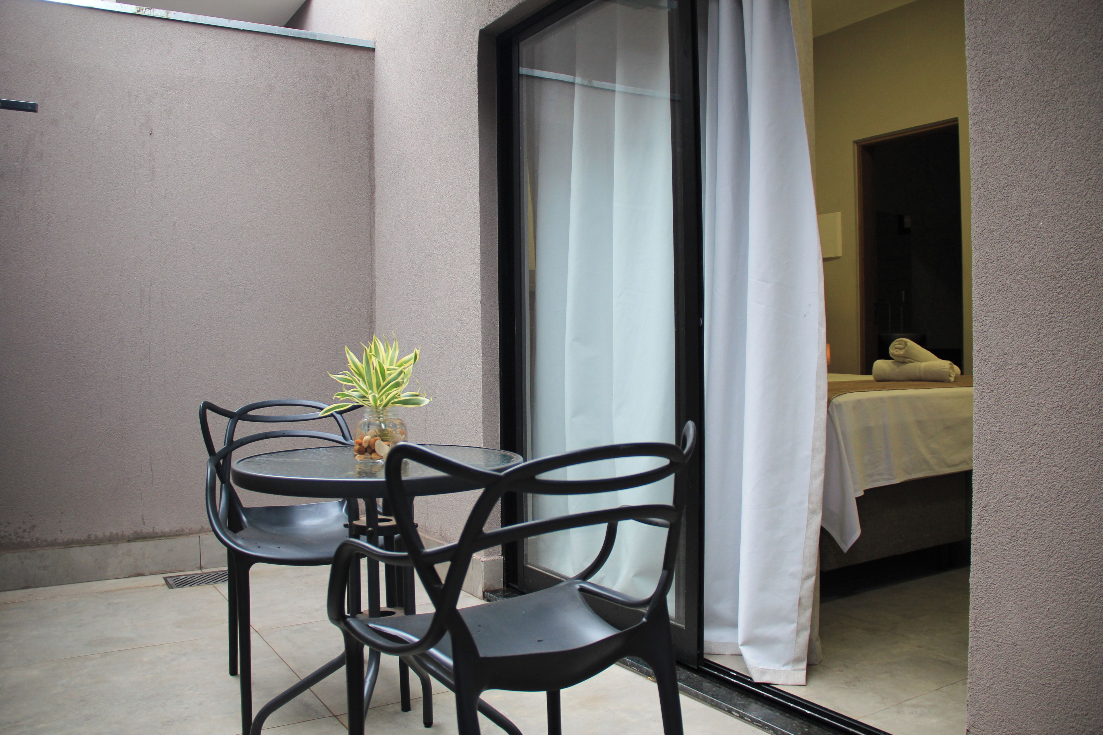 Un petit espace extérieur aménagé avec une table et des chaises, un pot de fleurs et une vue sur une pièce où se trouvent un lit et un rideau blanc.