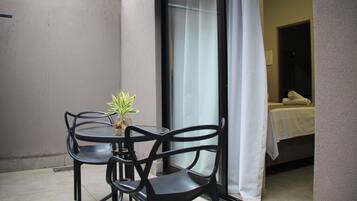 Un petit espace extérieur aménagé avec une table et des chaises, un pot de fleurs et une vue sur une pièce où se trouvent un lit et un rideau blanc.