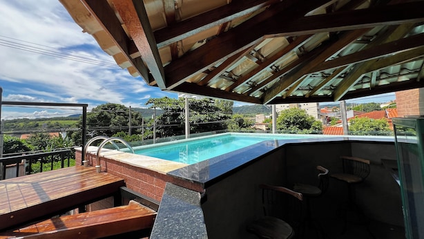 A rooftop pool with a wooden deck and bar area, surrounded by greenery and buildings in the background. - Pousada Casa Limão (Rifaina)