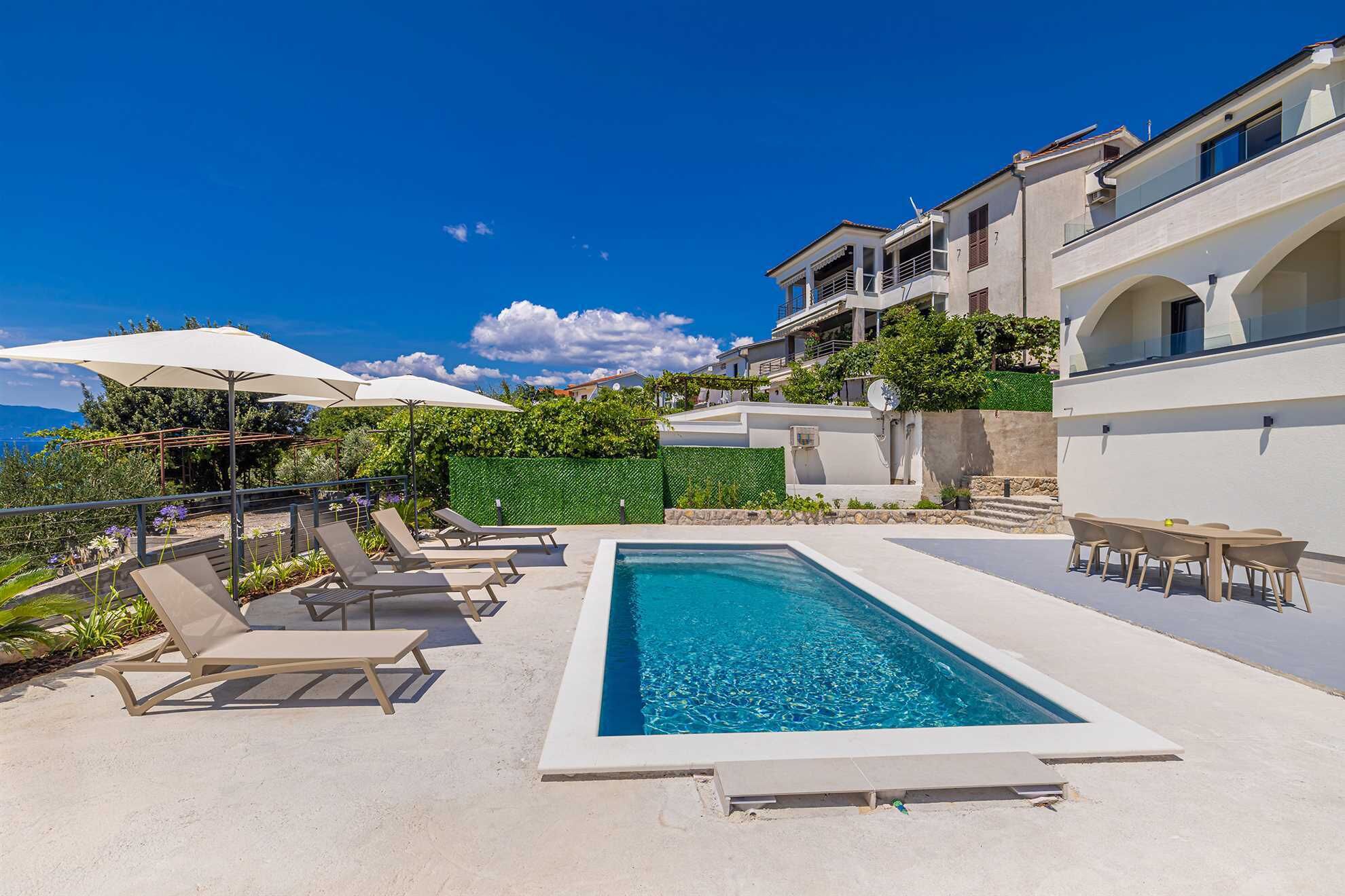 Une piscine rectangulaire entourée de chaises longues et de parasols, bordée d’un bâtiment blanc doté d’un balcon et offrant une vue sur la mer.