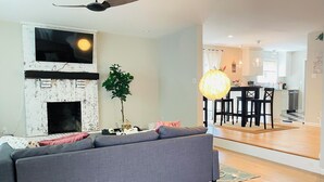 Un salon avec un canapé gris, une cheminée en briques blanches, un ventilateur de plafond et un coin repas comprenant une table et des chaises.