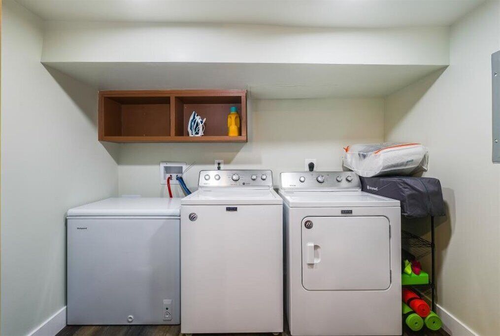 A laundry room with two front-loading washing machines and a dryer, a wooden shelf above with laundry supplies, and a small storage area with colorful items.