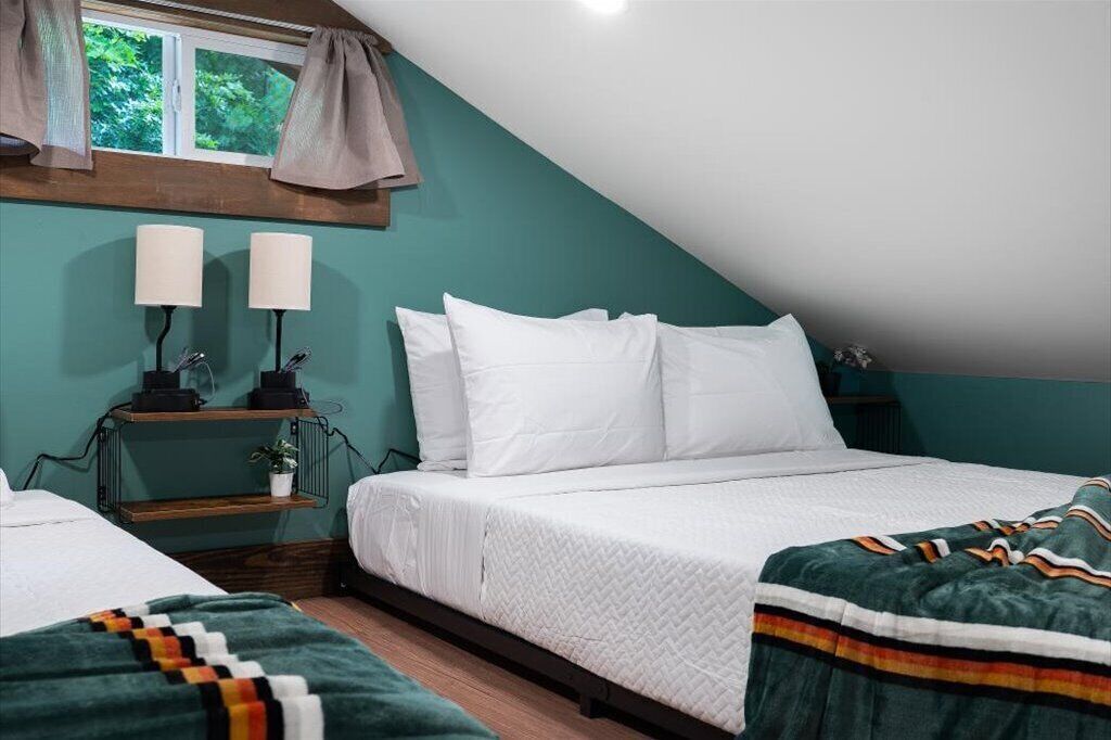 A bedroom with a sloped ceiling, a bed with white bedding, a bedside table with lamps, and a window with curtains.