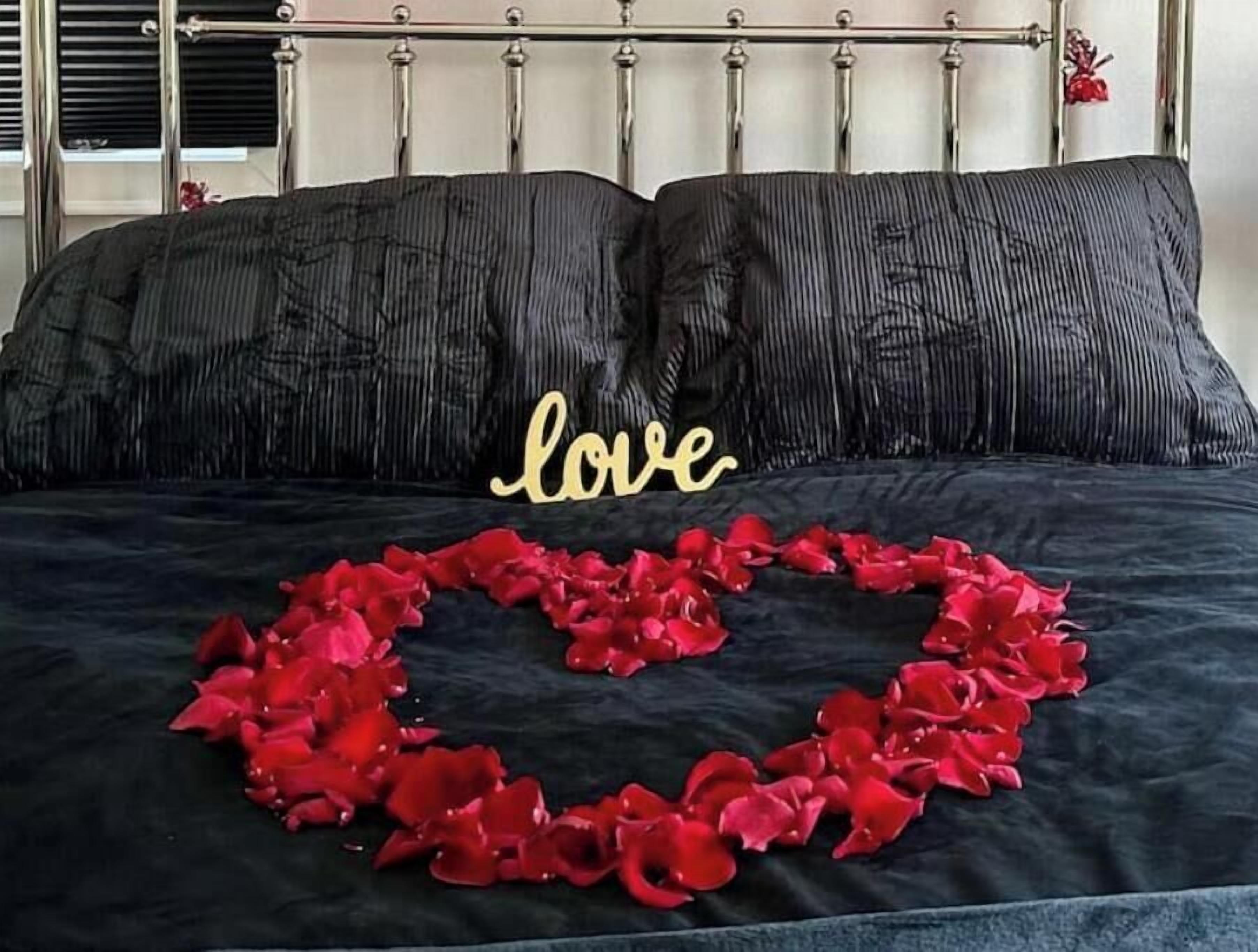 A bed with black bedding, a heart-shaped arrangement of red rose petals, and a 'love' sign in gold letters.