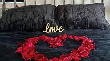 A bed with black bedding, a heart-shaped arrangement of red rose petals, and a 'love' sign in gold letters.
