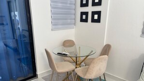A modern dining area with a glass table and four chairs, a window with blinds, and a wall with framed pictures.