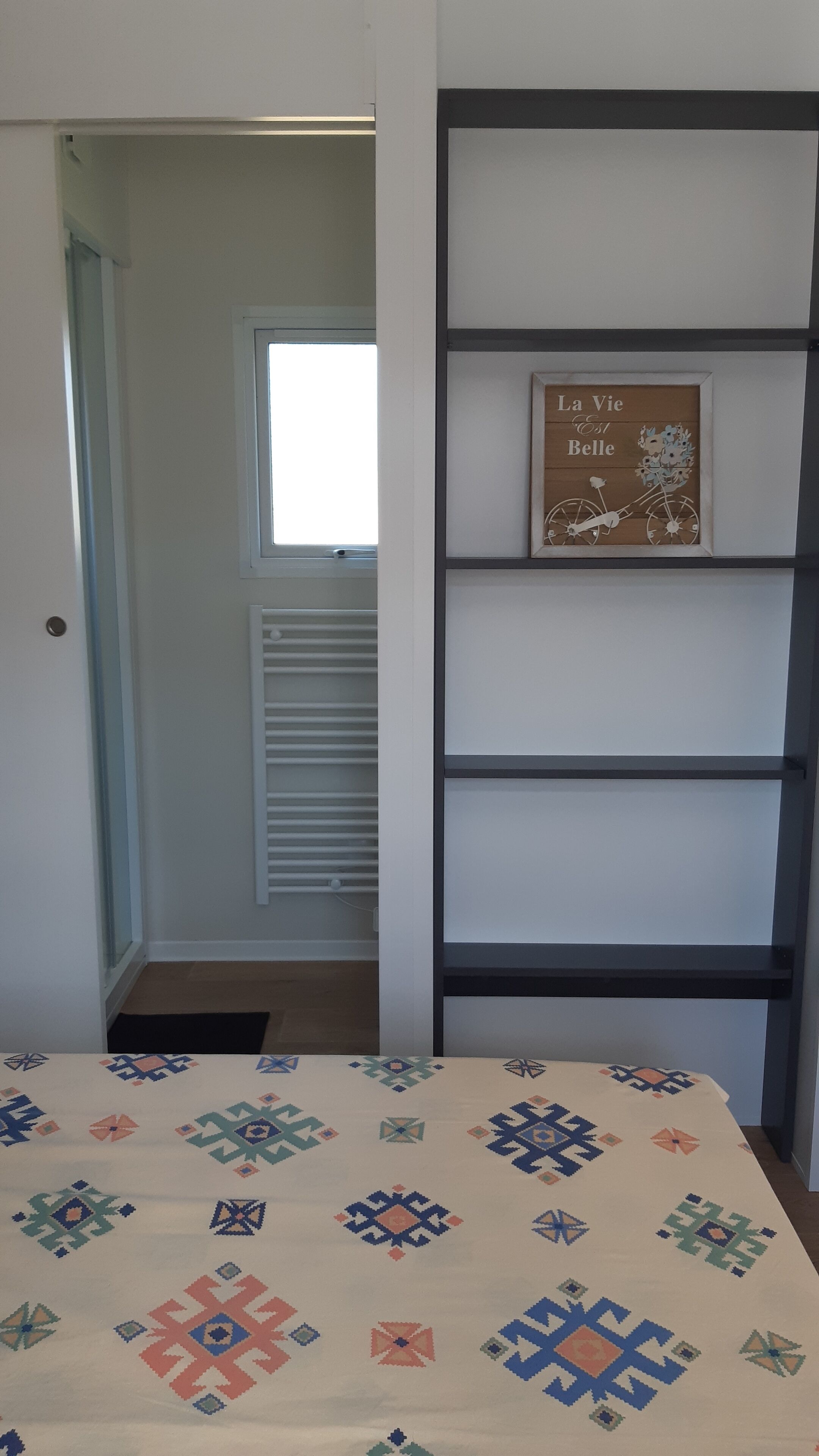 Une pièce avec un lit, une fenêtre, une porte et une étagère sur laquelle est posée une photo encadrée.