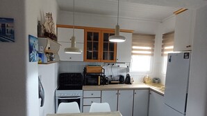 A compact kitchen with white cabinets, a black stove, a microwave, and a refrigerator. There are two white chairs at a wooden table, and a small ship model on the wall. - 100 meters from the blue flag awarded Mesi Beach. (Paralia Mesis)