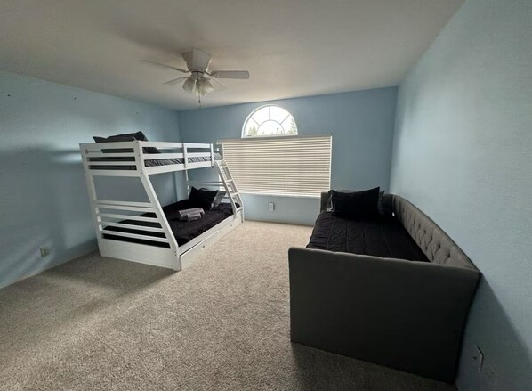 A room with a bunk bed, a single bed, a ceiling fan, and a window with blinds.