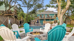 A backyard with a fire pit surrounded by turquoise and white Adirondack chairs, a wooden deck, and a house with a balcony.