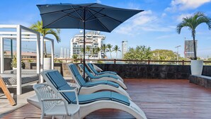 A rooftop lounge area with sun loungers and umbrellas, overlooking a cityscape with high-rise buildings. - 3rd-Floor Gem w/ Cruise Views & Rooftop Lounge (Puerto Vallarta)
